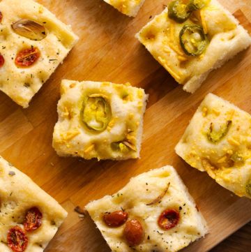 Square slices of jalapeno cheese focacccia and tomato herb focaccia on a wood cutting board.